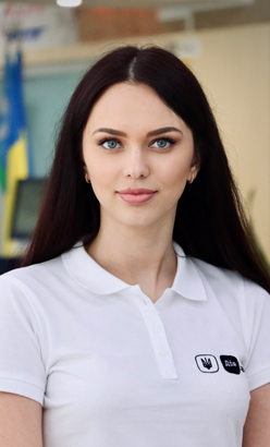 Жінка з темно-каштановим волоссям у футболці, у приміщенні, дивиться в камеру з упевненим виразом.